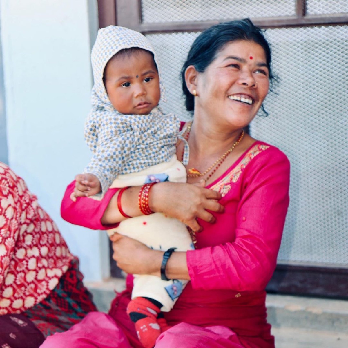 A grandmother holds her grandchild on the porch of their home in Indrawati, neighbours seated behind.