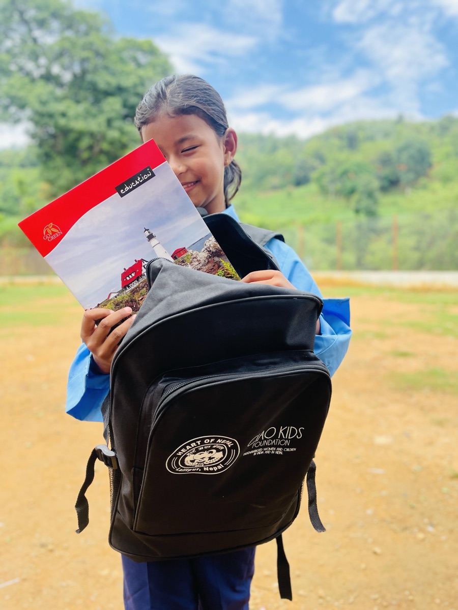 A scholarship student opens her Heart of Nepal x CIAO KIDS school bag — notebook in hand, hillside behind her.